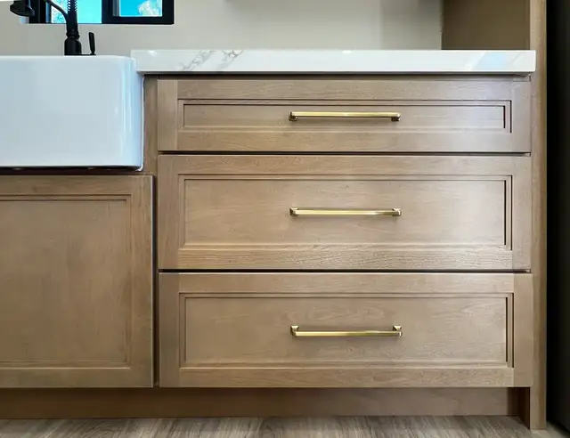 Close-up of stained wood shaker drawers with brass bar pulls beside a white farmhouse sink