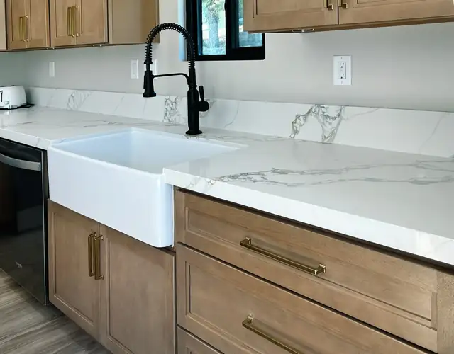 White farmhouse sink set in stained wood cabinets with brass pulls, a marble-look quartz top and a black faucet